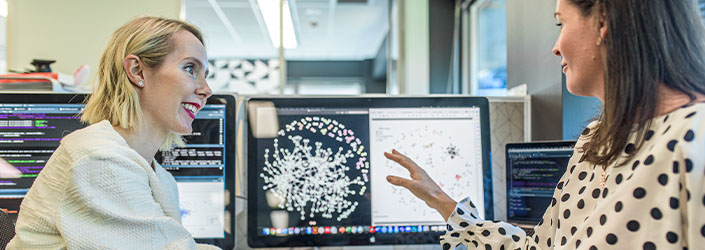 Two women speaking in front of a computer screen.