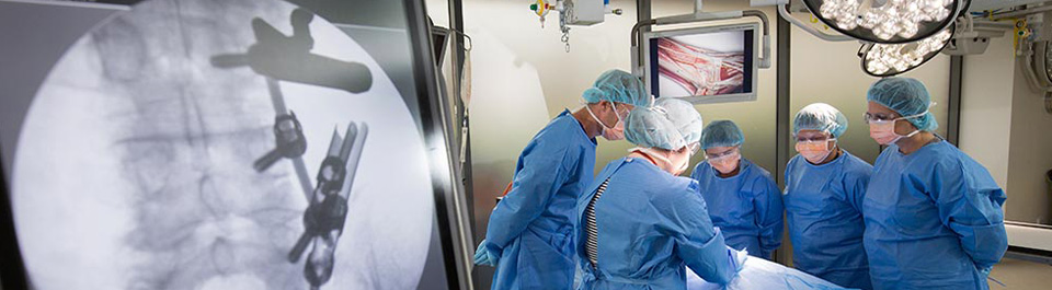 A group of people in an operating room dressed in scrubs, with grey screen showing the operation.