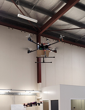 Two drones flying inside a warehouse, the higher one carrying a cardboard box.