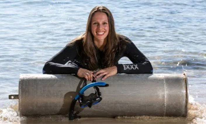 Dr Vanessa Pirotta posing behind a metal cylinder at the beach shore and wearing diving gear.